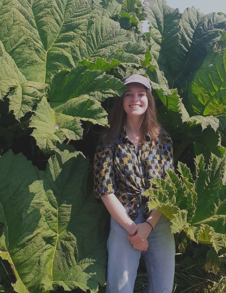 A picture of Isabel smiling, she's wearing a shirt with shirts on and is surrounded by very large leaves.